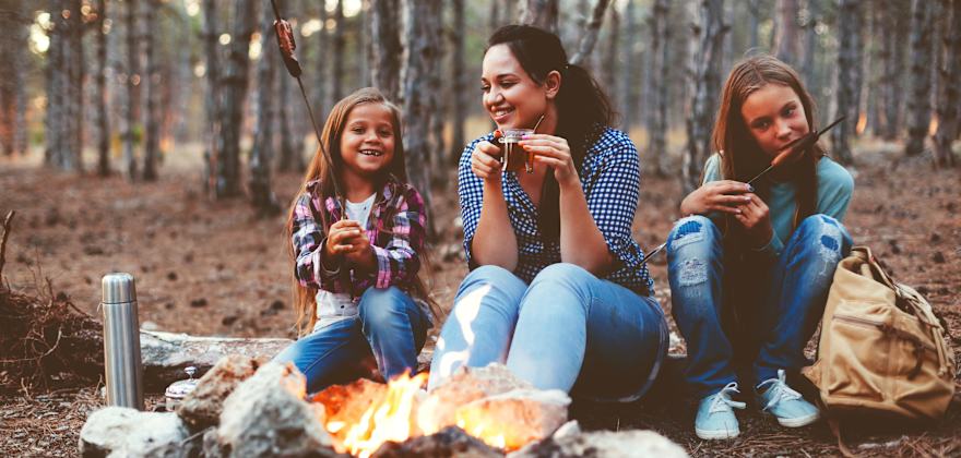 A mom and her two daughters sit around the campfire at a campsite in fall.