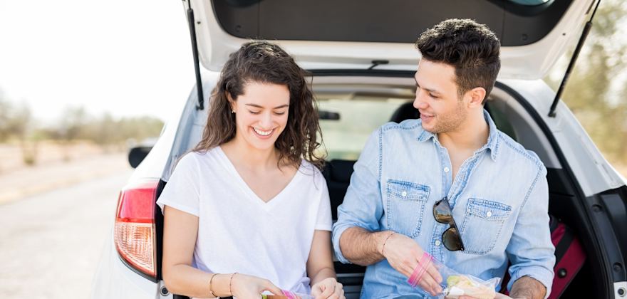 A couple sit on the tailgate and at a sandwich on the side of the road.