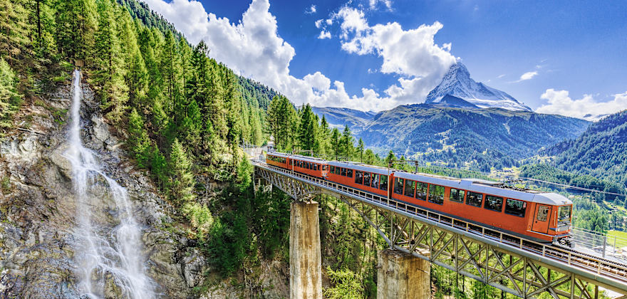A train crosses a bridge by a waterfall in the Swiss Alps.