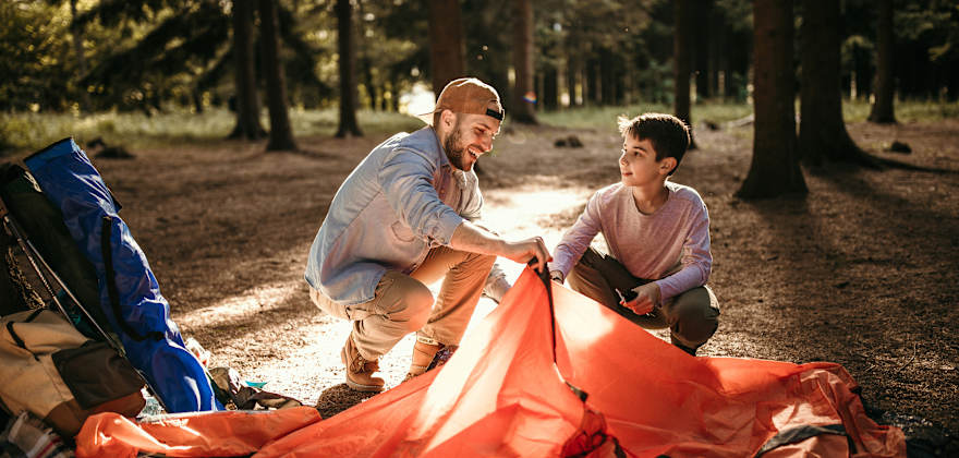 A dad and his son set up their tent at a campground  under the trees.