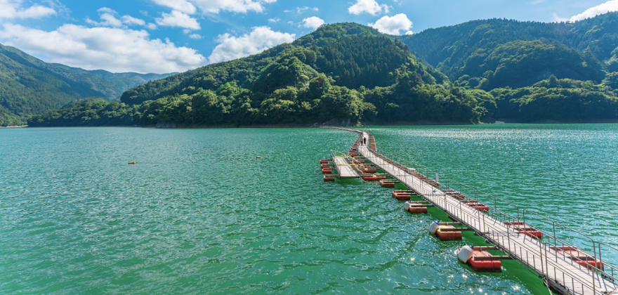 A few people walk along a floating foot bridge across the clear blue waters of Lake Okutama in Japan.