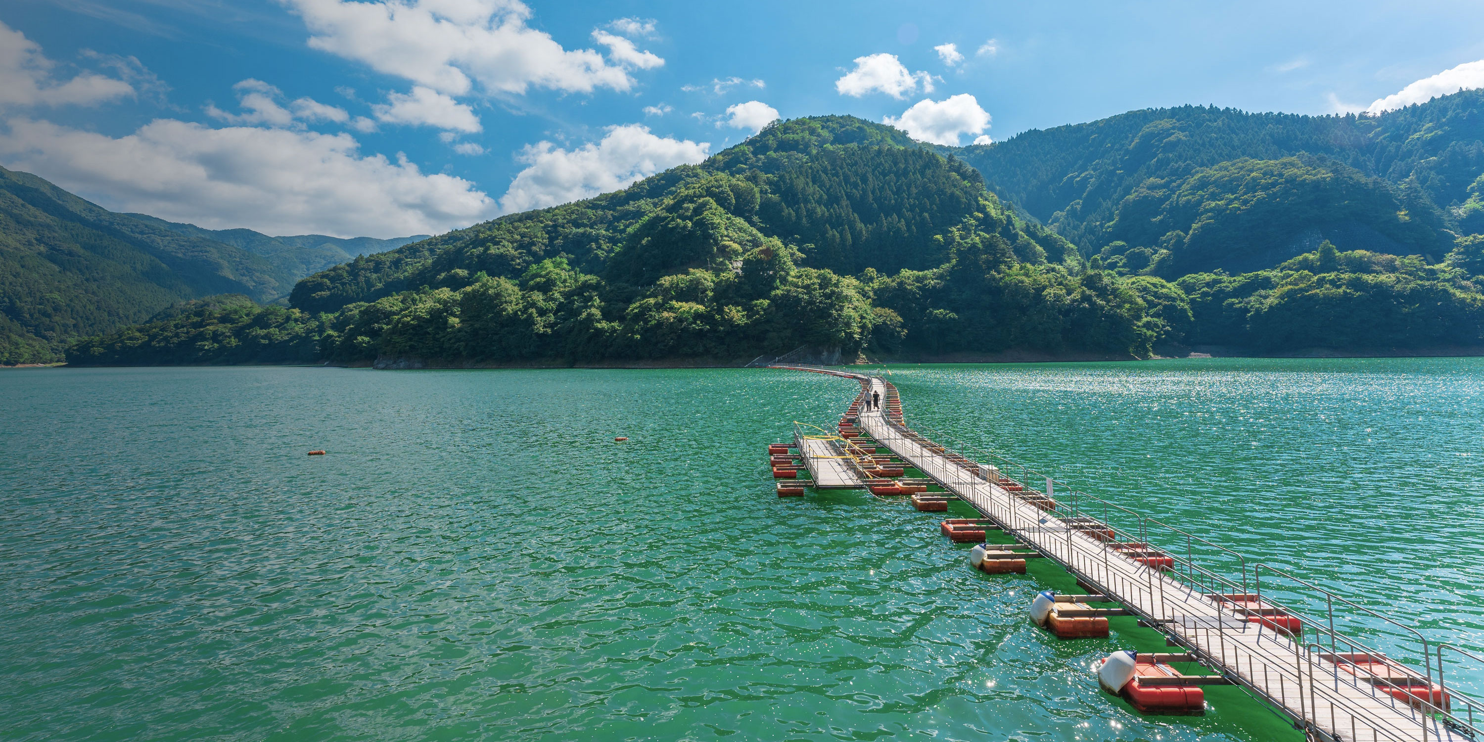 A few people walk along a floating foot bridge across the clear blue waters of Lake Okutama in Japan.