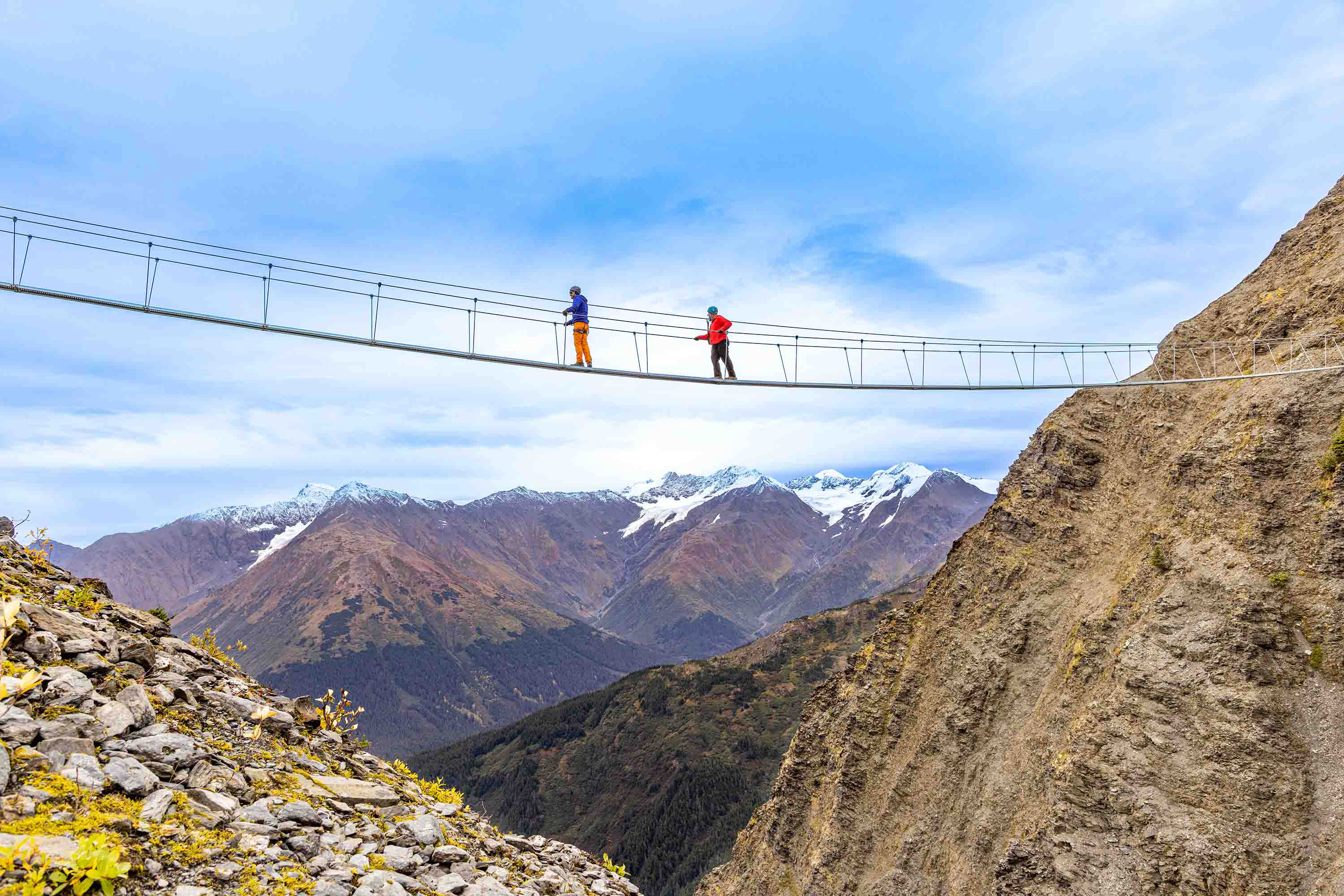 Two hikers navigate a cable bridge that runs between the peaks of Alyeska Mountain.