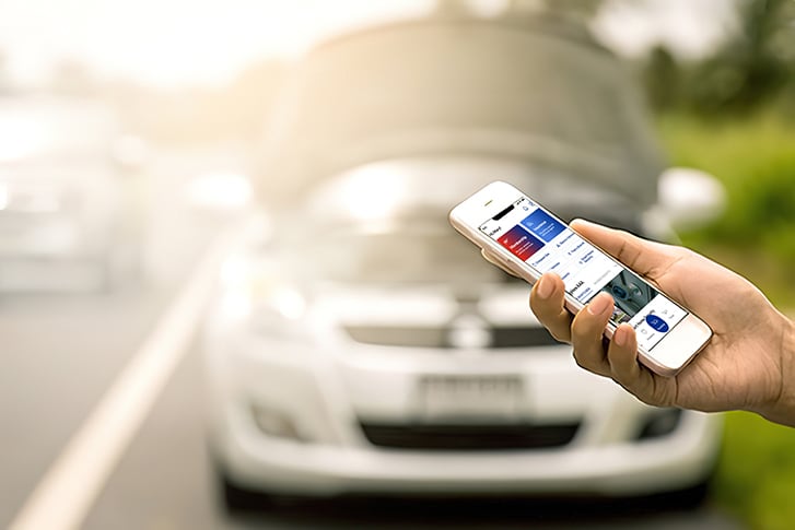 Hand of a AAA Member using the AAA app on their cellphone to call for roadside assistance, with broken down car in the background.