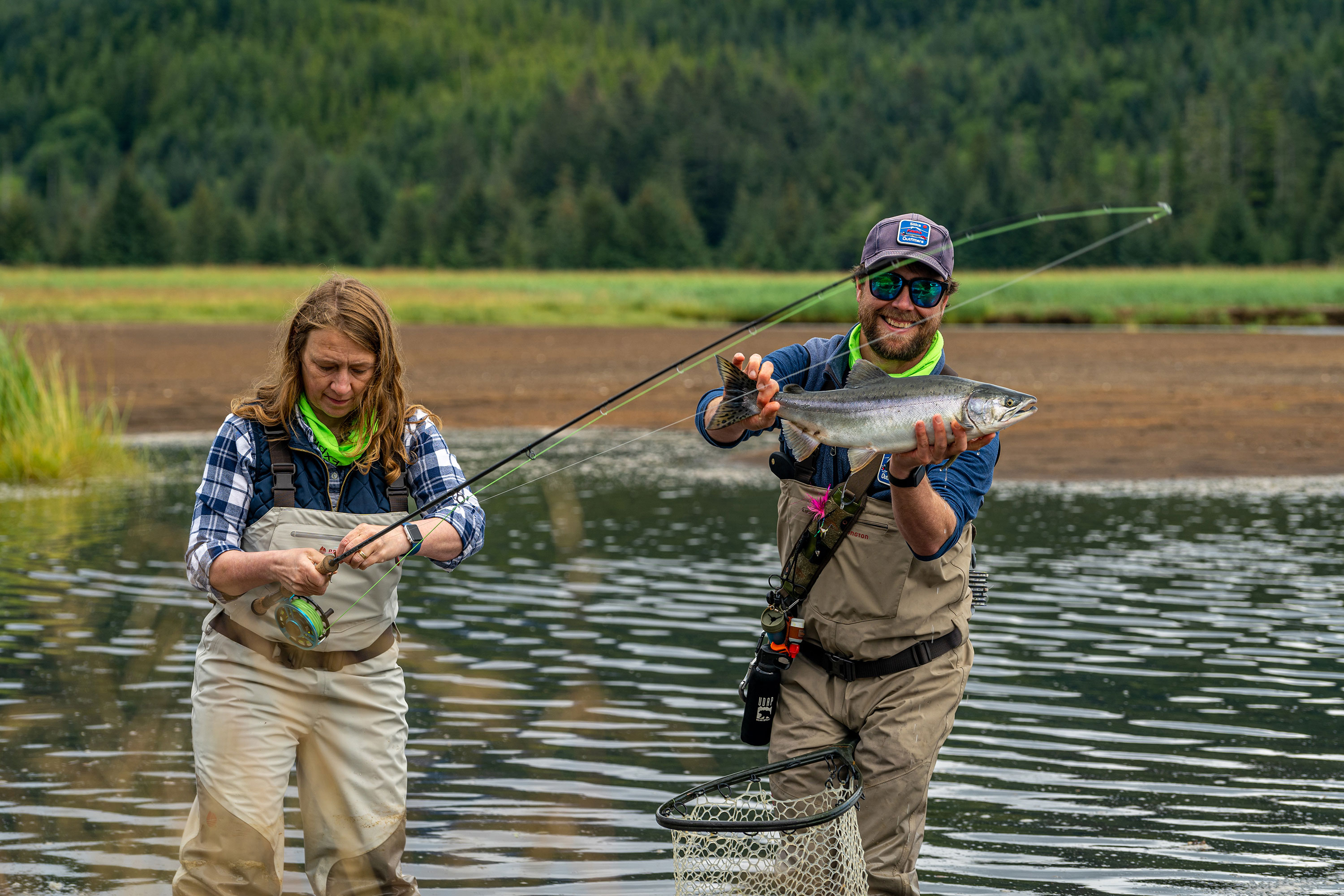 Two fishermen wearing waders stand in a river, holding a fish caught on a line.