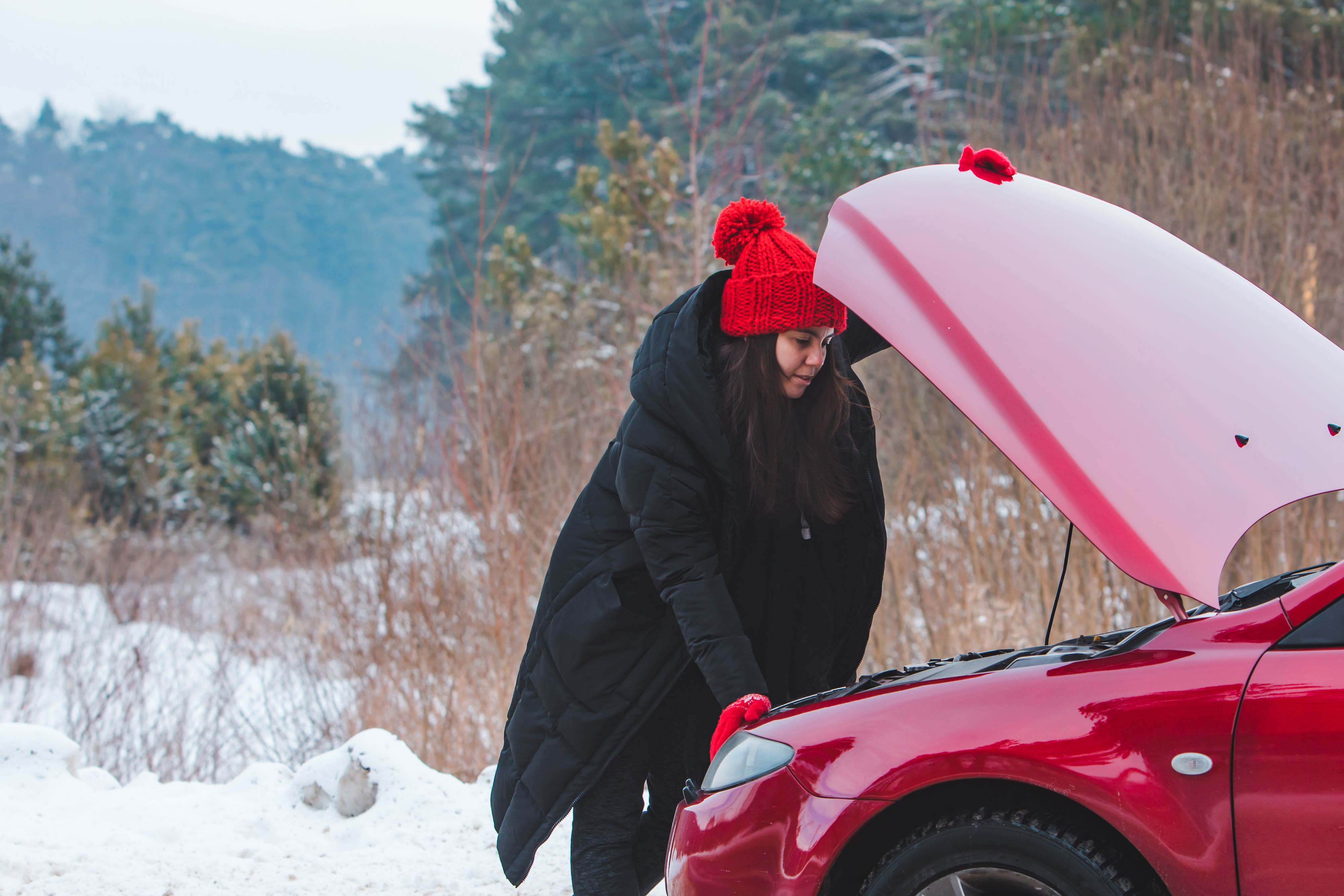 A woman in a coat and hat looks under the hood of her red car.