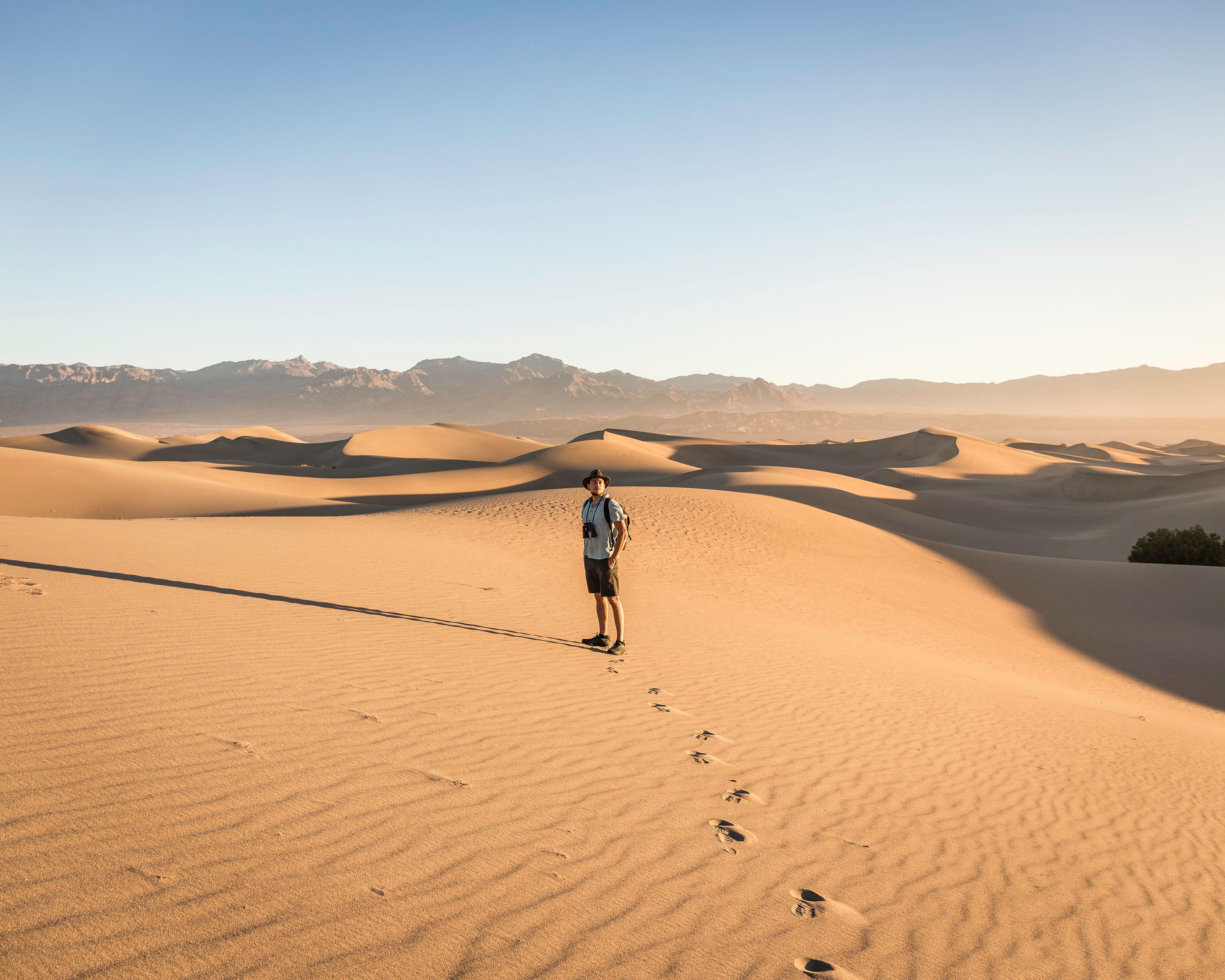 A man in shorts, t-shirt and sunhat walks through scenic sand dunes in Death Valley National Park, California