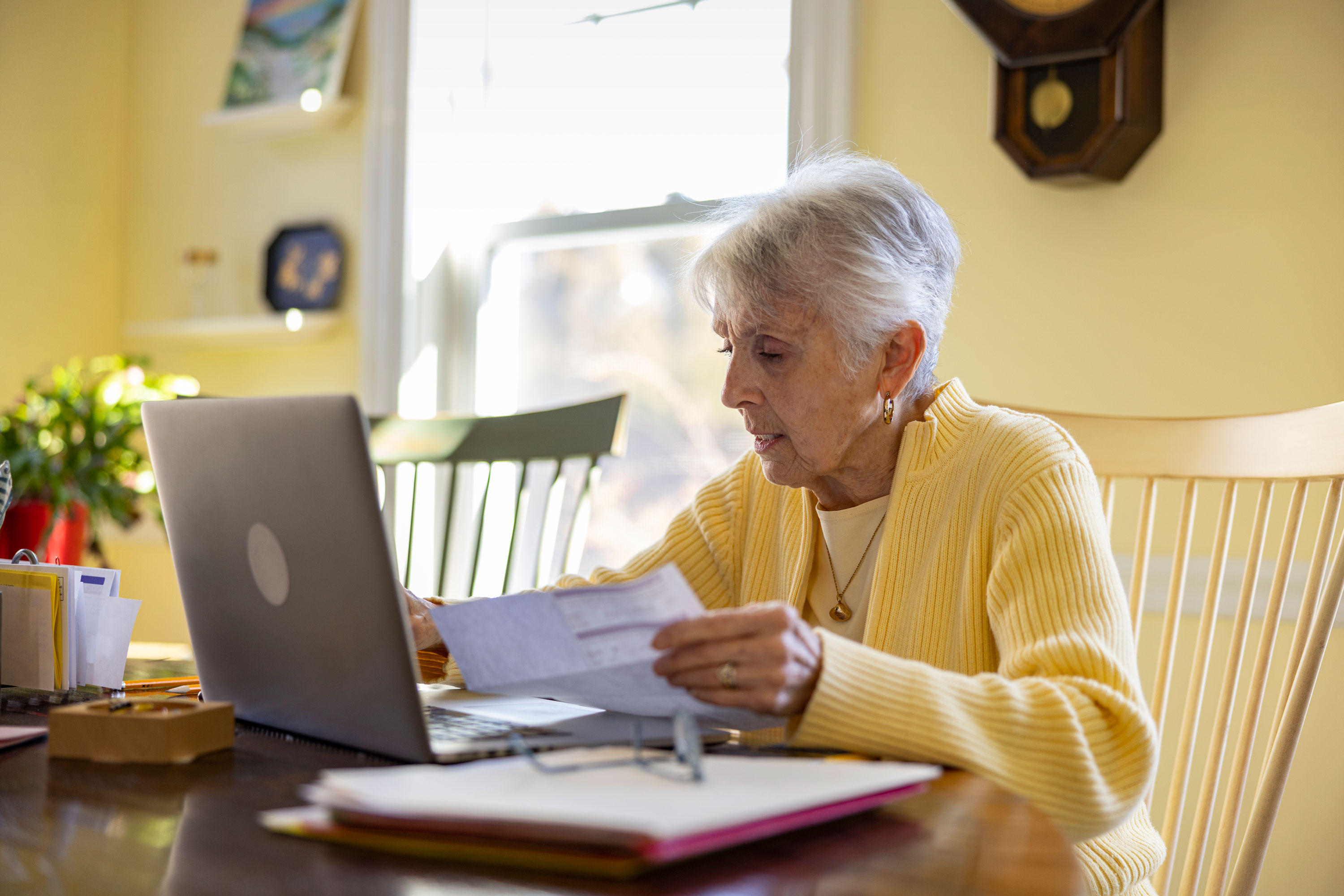 An older woman looks at financial statements online and on paper at her kitchen table.